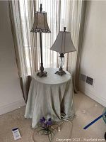 Overall view of two table lamps on round side table covered with beige tablecloth, floral arrangement on floor nearby.