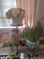 Wide shot of large ceramic planter with artificial bamboo, white metal planter with faux flowers on dark wood plant stand, glass vase with faux flowers on floor