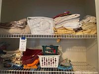 Shelving unit with various folded linens including bed linens, hand towels, and table linens, plus a clear packaged queen size mattress pad