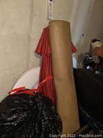 Photo of rolled up outdoor rug and folded red umbrella leaning against wall with black garbage bags nearby.