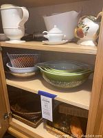 Photo showing white pitcher, decorative ceramic gravy boat with floral design, white ceramic sugar bowl with lid and saucer, metal wire basket, green Pyrex mixing bowls with floral pattern, wicker serving baskets, glass baking dishes, and kitchen utility items stored in wooden cabinet.