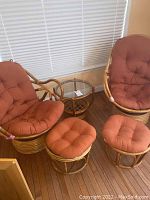 Two swivel rattan chairs with tufted orange cushions, two matching ottomans with cushions, and a round glass top side table in a corner with wood flooring and window blinds.