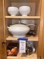 Shelf with large white soup tureen with lid and ladle, four white soup bowls on upper shelf, brown soup crock with lid and handle on bottom shelf, Garlic Baker box, white measuring cups and funnels also visible