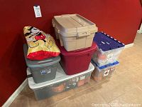Six plastic storage bins and one Mickey Mouse bag stacked in a corner, containing fall and holiday decor.