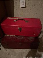 Red metal tool box placed on top of a dirtier dark metal tool box in a basement setting, showing overall condition and labels on boxes.
