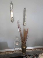 Pair of narrow vertical mirrors on wall above white metal stand and brass-tone vase with dried fillers