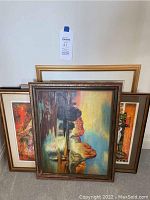 Multiple framed wall art pieces stacked against a wall including two landscape paintings, a colorful art piece, and an empty gold frame