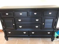 Front view of large black wooden dresser showing eight drawers, two shutter-style storage doors, and stainless steel knobs. Minor surface wear and scratches visible.