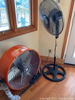 Holmes oscillating stand fan and large orange construction fan on hardwood floor near door and window.