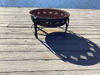Fire pit sitting on wooden dock near water with visible shadows. Shows circular metal bowl with diamond cutouts and rusty grate on top.
