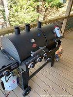 Full view of dual-chamber grill and smoker with side burner, propane tank, charcoal bag, and lighter fluid on deck