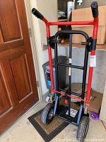 Front view of red and black Milwaukee hand truck standing upright indoors with shelves and items behind it.