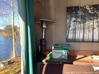 Photo of propane heater with metal reflector top and control knob near wall and patio curtain, includes box with firelogs on tile counter