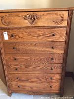 Front view of the tall oak chest of drawers showcasing carved floral design on top drawer and six lower drawers with round knobs.