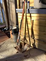 Photo showing one metal shovel with wooden handle and two garden rakes with wooden handles and metal heads leaning against a wooden structure outdoors.