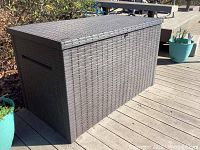 Front and side view of rectangular brown faux wicker outdoor storage box sitting on dock planks with green plants near it