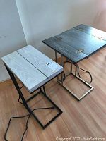 Pair of Tainoki folding end tables in light and dark wood finishes with metal bases, visible power cords on wooden floor.