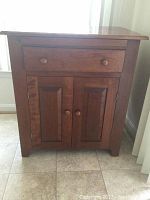 Front view of medium brown wooden cabinet with one drawer and two doors, showing knobs and scratches on the surface.
