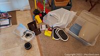 Photo shows various cleaning tools on floor including two dust mops, two toilet plungers, a wicker basket, plastic bins, flashlights, and other small items scattered on carpet and tile floor.