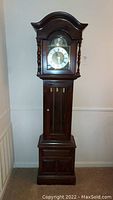 Full front view of the tall wooden grandfather clock showing the clock face, pendulum, weights, and cabinet base.