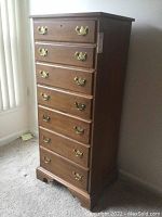 Photo showing full view of the tall wooden lingerie chest with seven drawers and brass handles. Chest is medium brown color with classic Ethan Allen styling, set against a light carpeted floor.