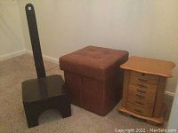 Photo showing the black stool with a tall backrest and handle cutout, brown tufted storage ottoman, and wooden jewelry box with multiple drawers.