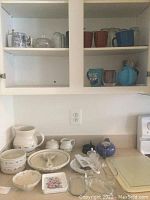 White and brown ceramic mugs on upper cabinet shelves and various glass bowls and butter dishes on countertop below.