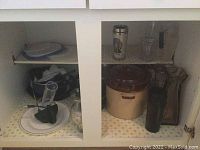 White kitchen cabinet interior with glass measuring cups, coffee mug, ceramic pots, plastic lids, plastic plate, plastic dog treat containers, and kitchen utensils.