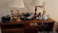 Photo of a wooden cabinet top with four table lamps with various shades, wood wreath with artificial flowers, phones, Emerson alarm clock, candle, and white dove flower pot.