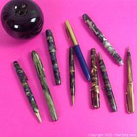 Nine vintage pens and pencils of various brands laid on bright pink background with a green apple for size reference