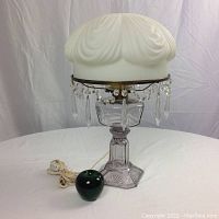 Full view of the antique electrified lamp with green apple for size reference showing glass base, milk white porcelain shade with brass and hanging crystals.
