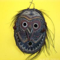 Front view of shaman mask showing painted facial features including large circular eyes and beak nose, with hair-like fibers tied to edges.