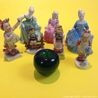 Seven porcelain figurines arranged on yellow background including three Royal Doulton figures and four Hummel figures with a green glass apple for scale.