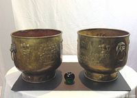 Front view of two large antique French brass jardinieres with a 3-inch glass apple for size reference, showing hammered brass exterior with lion-faced ring handles and French mythological embossing.