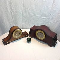 Two wooden mantel clocks displayed side by side on a white surface, with a green glass apple between for size reference.