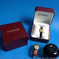 Two watches displayed on blue background with red Wittnauer box and green glass apple for scale