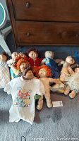 Photo showing nine Cabbage Patch Dolls lying on floor, assorted outfits and hair colors, dolls about 15 inches tall. Visible wear and staining on dolls and clothing.