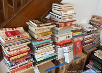 Photo showing large stacks of assorted cookbooks, both hardcover and paperback, piled on boxes and a table near stairs.
