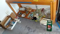 Wide view showing wooden shelf holding glassware, craft items, and iron beneath with some items on floor near box.