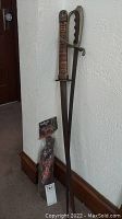 Full view of sword leaning against wall with carrying strap in package at foot