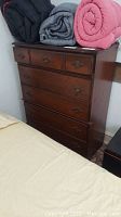 Dark wood dresser with six drawers, two smaller top drawers and four larger bottom drawers, visible wear including scratches and scuffs shown. Thick bedding stacked on top, beige bed visible in foreground.
