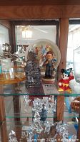 Shelf display showing Mickey Mouse and Pinocchio figurines, film camera figurine, round decorated plate, and glassware pieces behind