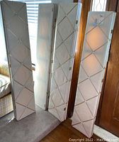 Four ivory fabric room dividers standing folded in room near window and door. Dividers have diamond pattern created by crossing fabric strips on front.