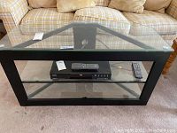 Front view of media stand showing glass top, middle glass shelf, and black metal frame with DVDs and remotes on the shelf (not included in lot).