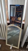 Two mirrors leaning against wall: larger rectangular black plastic framed mirror and smaller vintage arched painted wood framed mirror on floor.