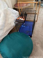 Black metal dog crate with blue base and black padded cushion inside, wooden sliding gate standing behind the crate, black dog harness sitting on top of crate, and large round green cushioned dog bed placed on floor.