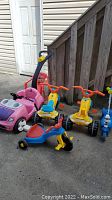 Wide shot showing two scoot along carts, one blue and yellow stand-up scooter, one pink pedal push car with black handle for pushing, and one red plastic chair on outdoor concrete.