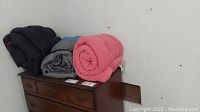 Three rolled sleeping bags sitting on a wooden dresser, colors black, gray, and pink visible.