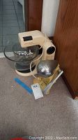 Vintage Sunbeam Mixmaster electric mixer with two glass mixing bowls and Metal Juice-O-Mat juicer with additional accessories and paper documents on floor next to wood cabinet.