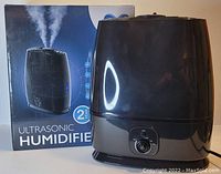 Black ultrasonic humidifier in front of box, showing the product and packaging, with control dial and LED indicator lights visible.